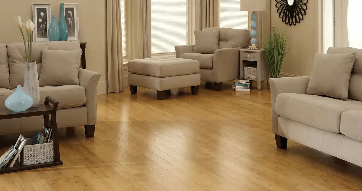 A living room with professionally installed bamboo flooring, beige sofas, a matching ottoman, a side table with a blue lamp, vases, framed pictures, and large windows with blinds and curtains in Minneapolis, MN